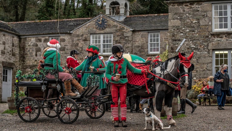 A horse and trap, decorated for Christmas with green tinsel, in front of grey stone building with a small clock tower. There are four people dressed as Christmas elves, holding takeaway cups of hot drinks. The person at the forefront is holding the horse's bridle and smiling at a small terrier sat by their feet. .